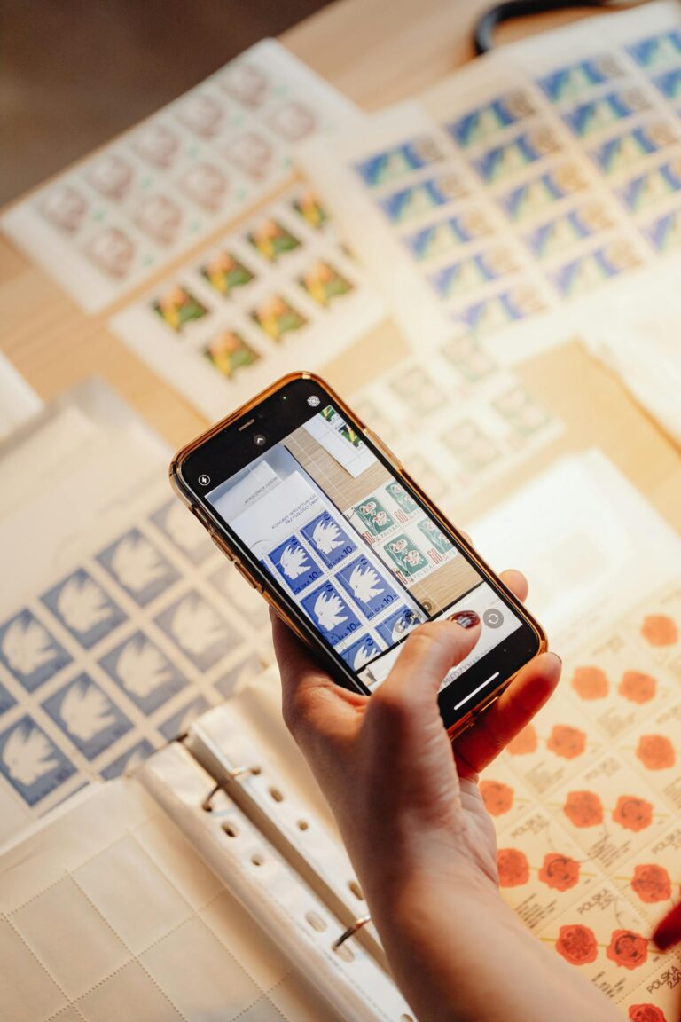 Close-up of hand holding smartphone photographing colorful stamp collection on table.